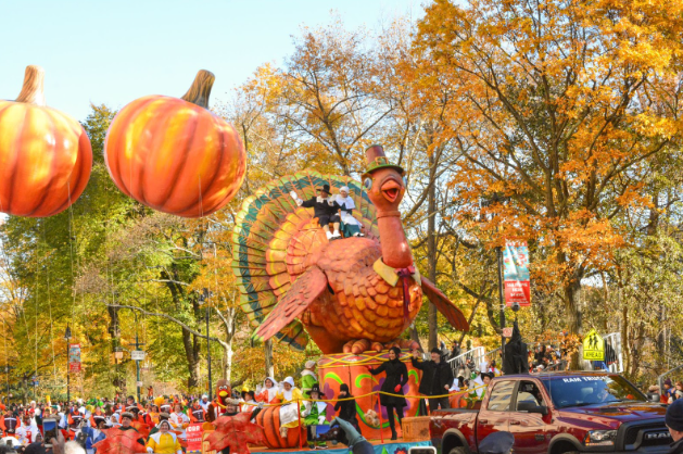 Photo Courtesy of TomH2323 - Macy's Thanksgiving Parade, CC BY 2.0, https://commons.wikimedia.org/w/index.php?curid=114069441. An image of the Macy’s Tom Turkey float. The float contains a giant orange turkey with people dressed up as pilgrims to celebrate Thanksgiving. 